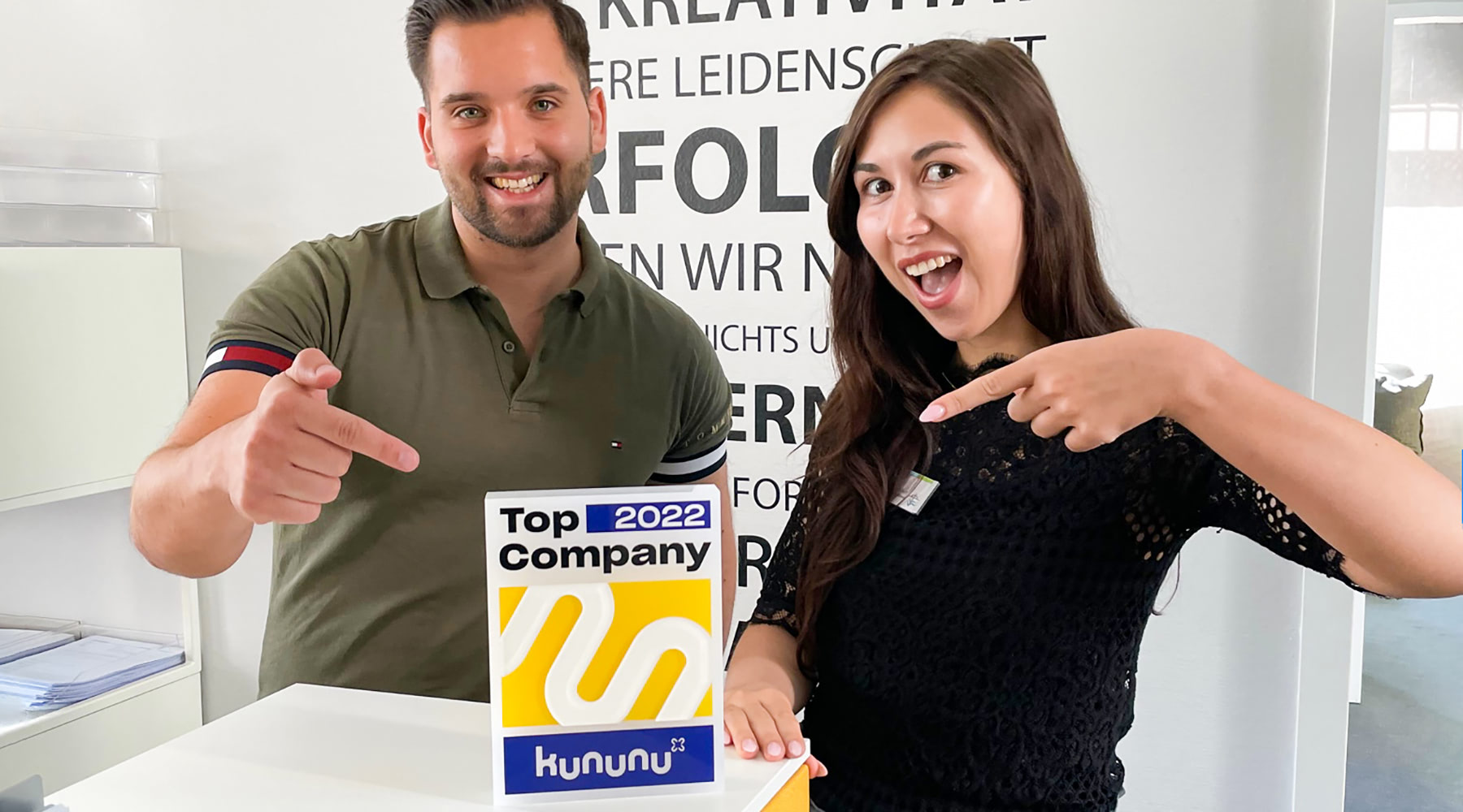 Leeroy Stiller und Charlotte Weber zeigen lächelnd auf die Auszeichnung mit dem Top Company Siegel 2022