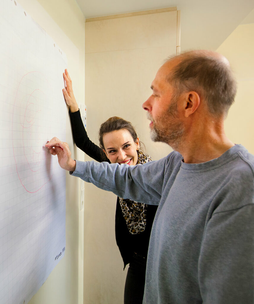 Ein Patient bei der Ergotherapie malt auf einem großen Blatt an der Wand und die Therapeutin schaut lächelnd aus dem Hintergrund heraus dabei zu