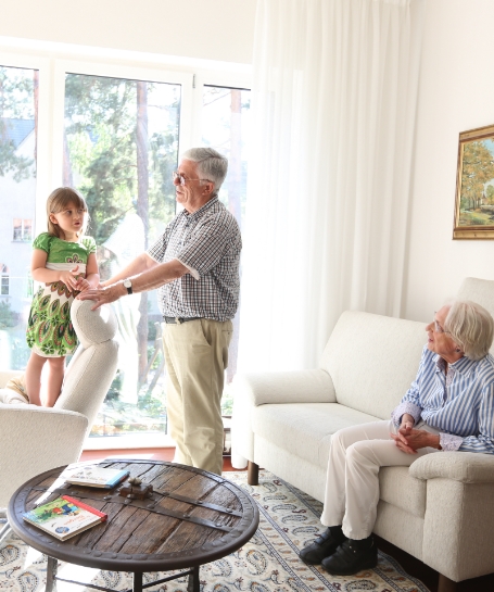 Ein kleines Mädchen steht vor einer großen Fensterfront auf einem Sessel, redet mit ihrem Großvater, der den Sessel festhält und die Großmutter schaut von der Couch aus zu ihnen